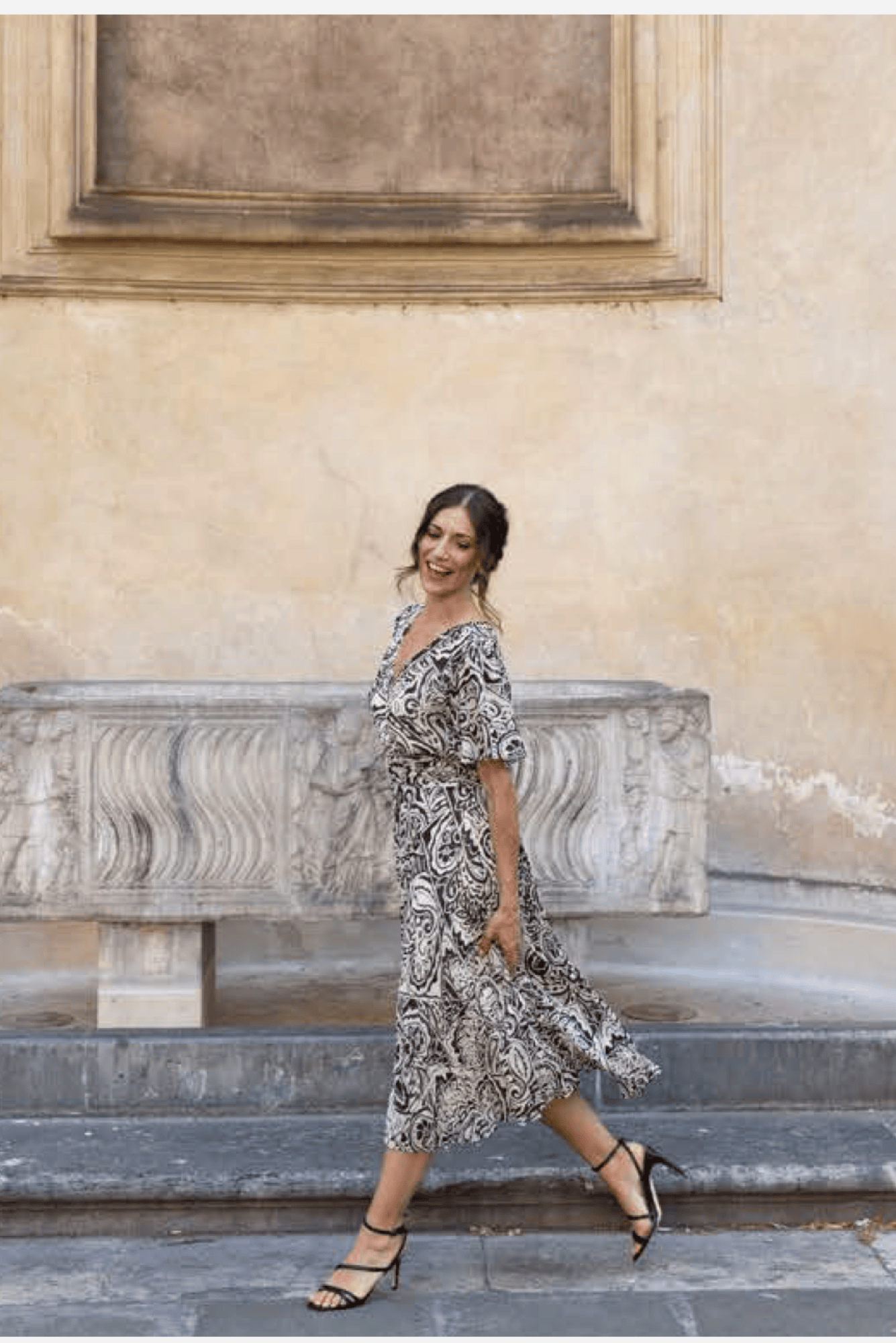 A woman wearing a black and ivory paisley midi dress walks down stone steps in front of a beige stucco wall.
