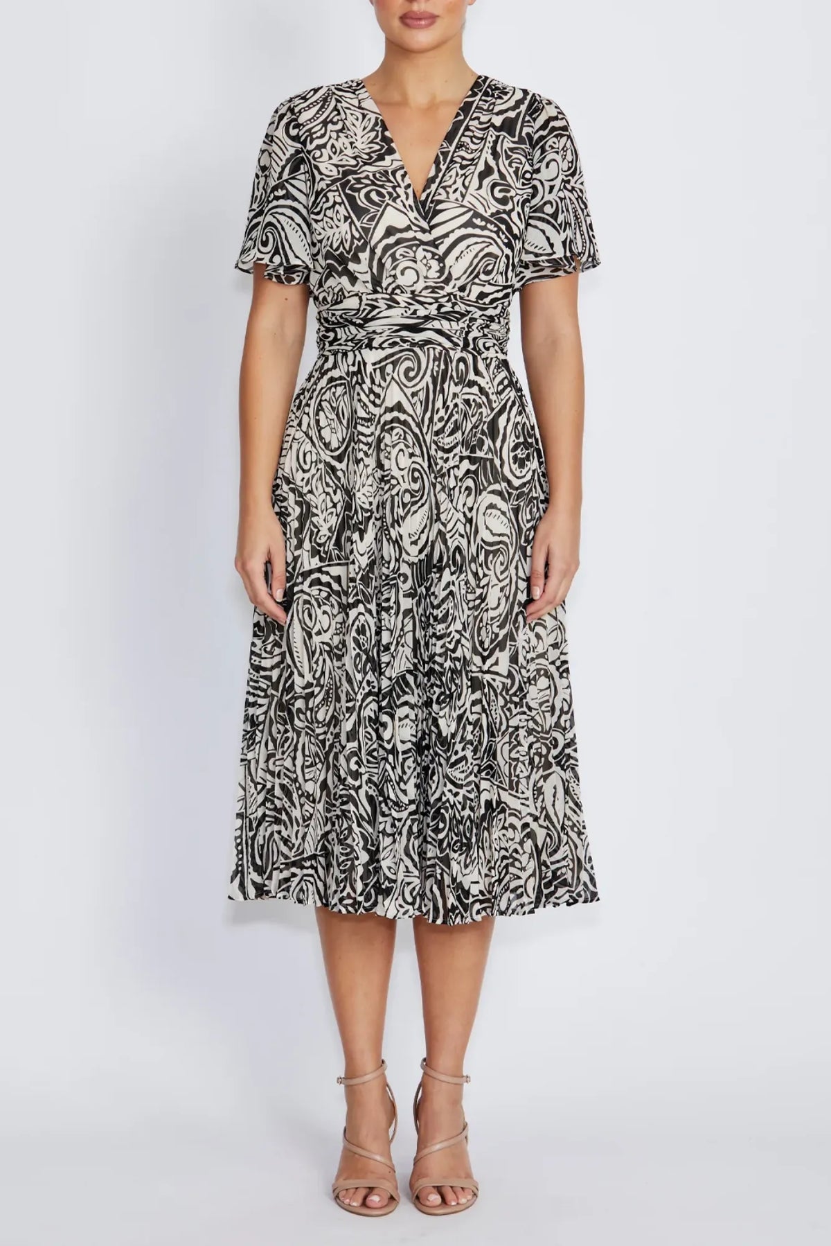 Front view of a woman modeling a flutter sleeve midi dress with a black and white paisley t pattern,  V-neckline, and a matching tie belt at the waist, paired with strappy high-heeled sandals against a light gray studio background.