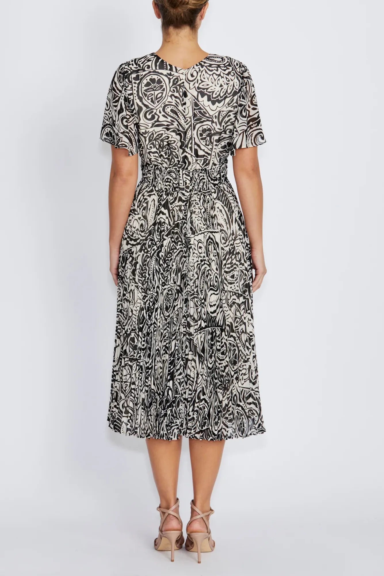 A woman viewed from the back wearing a flutter sleeve midi dress with an abstract black and white paisley pattern and beige strappy high-heeled sandals, set against a plain light gray studio background.