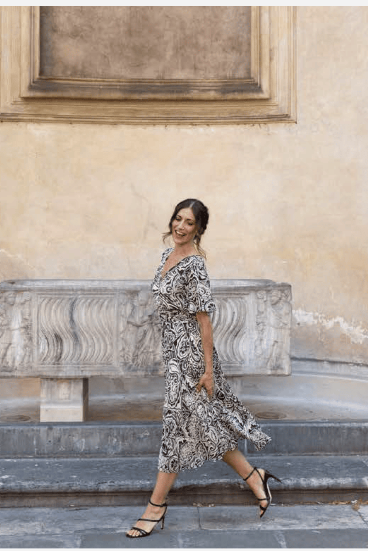 A woman wearing a black and ivory paisley midi dress walks down stone steps in front of a beige stucco wall.
