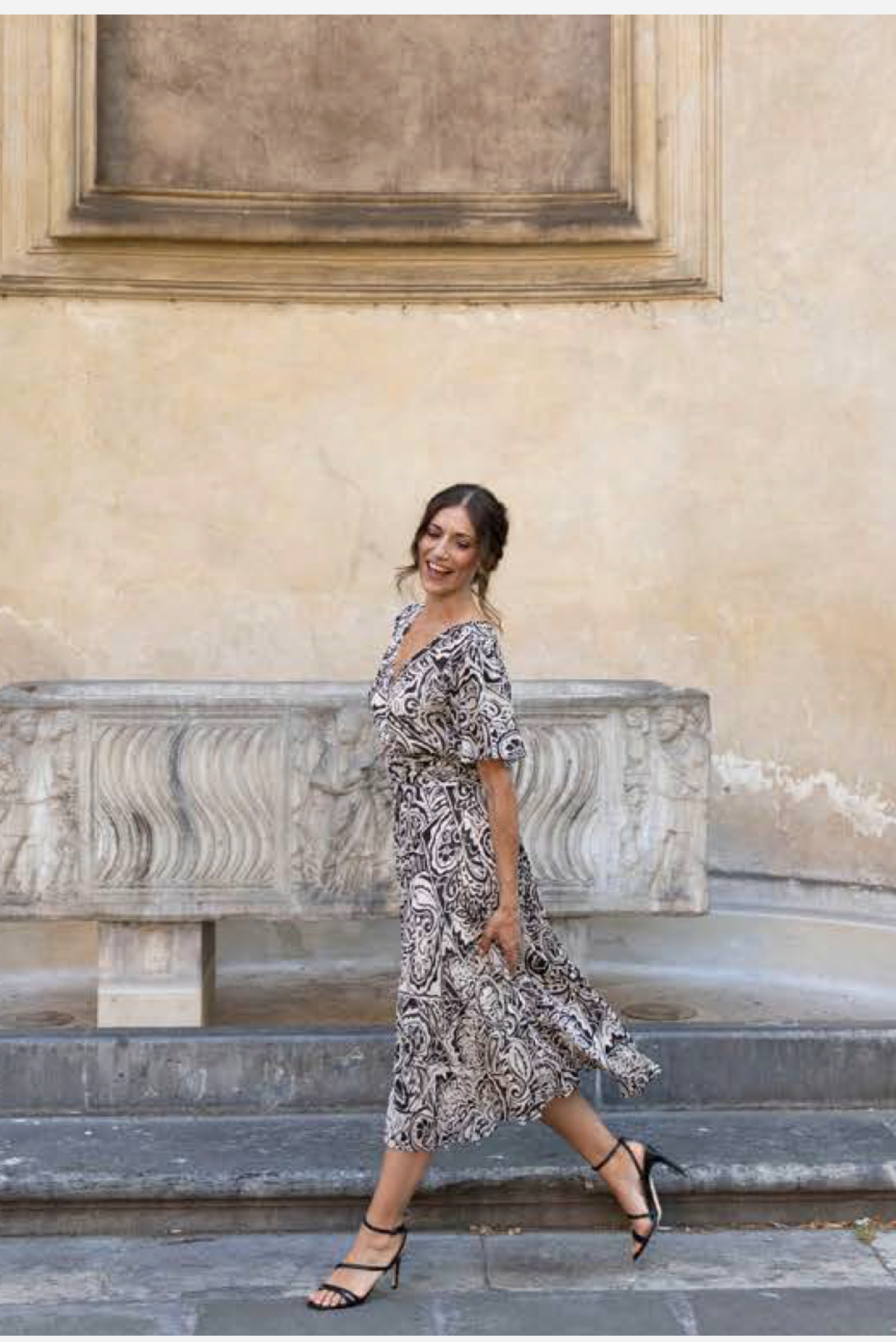 A woman wearing a black and ivory paisley midi dress walks down stone steps in front of a beige stucco wall.
